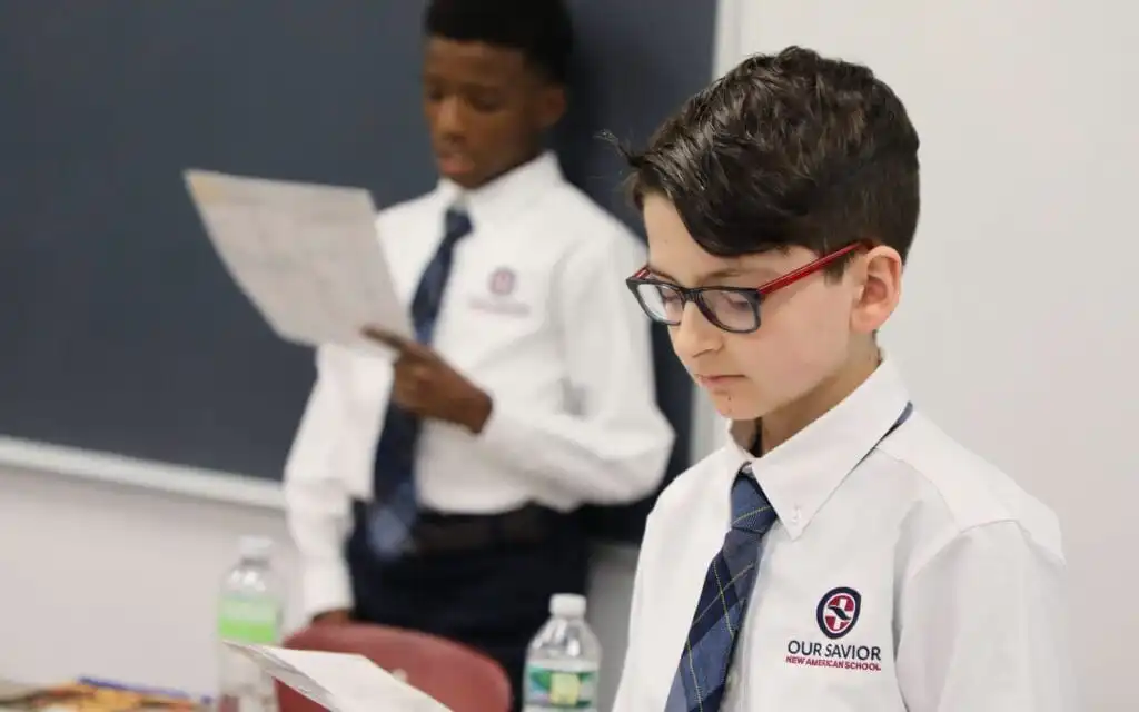 Two boys stand at the front of a classroom and read to the class.